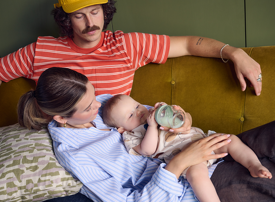 dad in striped top next to mum in shirt holding baby and glass bottle on mustard sofa