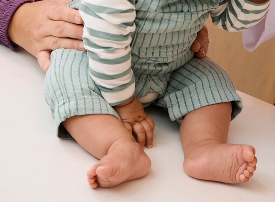 A seated baby wearing green-striped pants and a white striped shirt, with a hand resting on their leg.