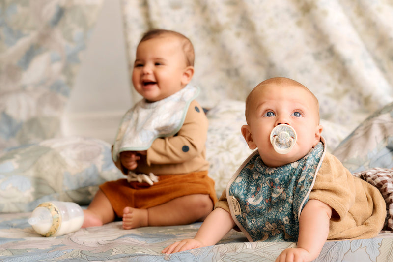 one baby smiling sitting up and another baby lying down with soother in mouth