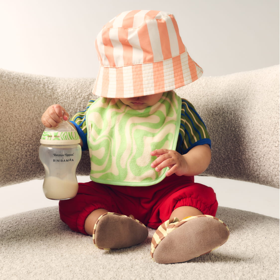 Baby sat on sofa wearing a summer hat using Tommee Tippee x Binibamba Natural Start Bottle and Bib