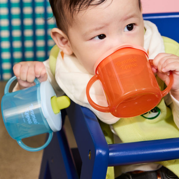 Toddler sitting in a highchair drinking from one Tommee Tippee first cup and holding another in the other hand