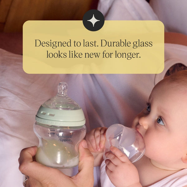 A parent sitting on bed with their baby and holding a Tommee Tippee Natural Start glass bottle with text stating 