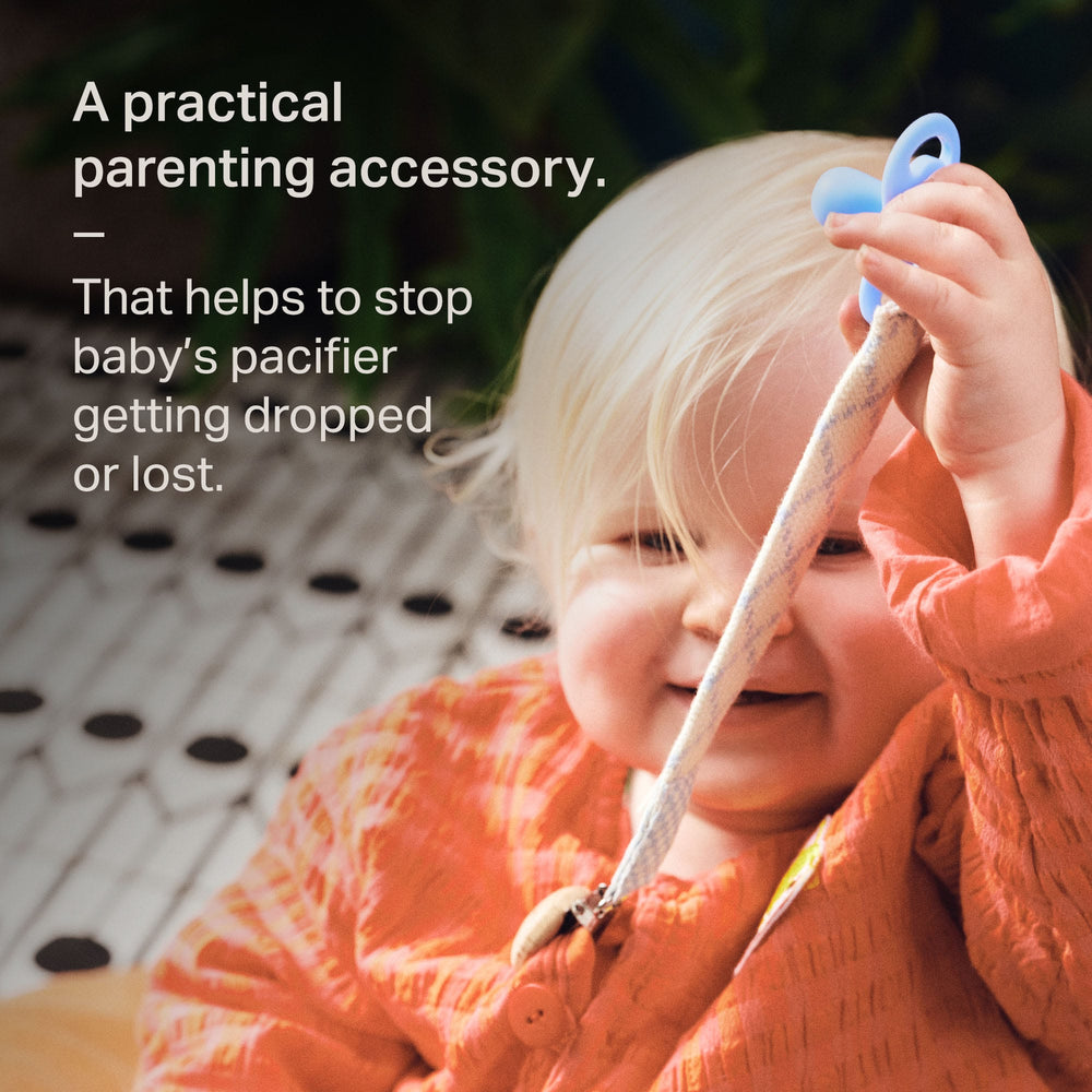 Pacfifier clip attached to baby's jacket while they hold their soother with text overlay.