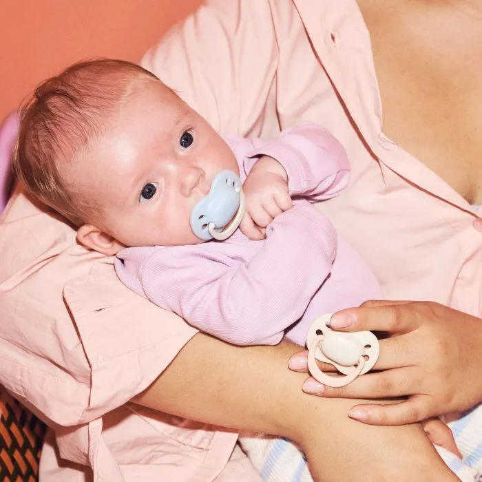 Parent cradling baby with a newborn pacifier while holding another newborn paci.