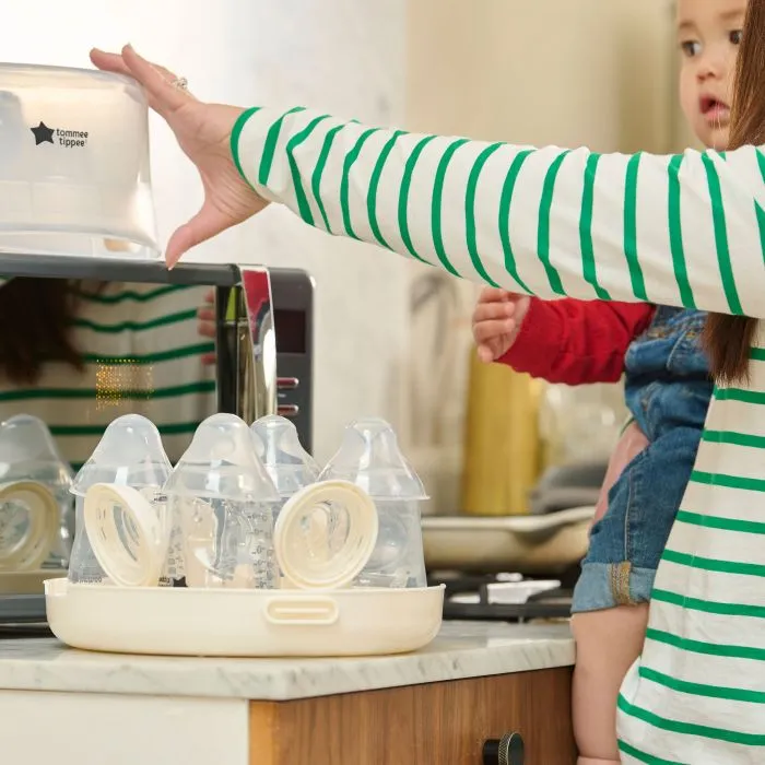 Woman holding a baby on her hip and filling a microwave steriliser ready to go in the microwave