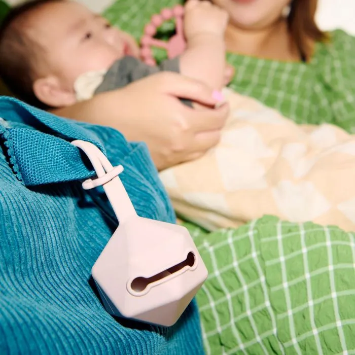 Pink soother carry case attached to bag strap with baby holding teething ring in the background.