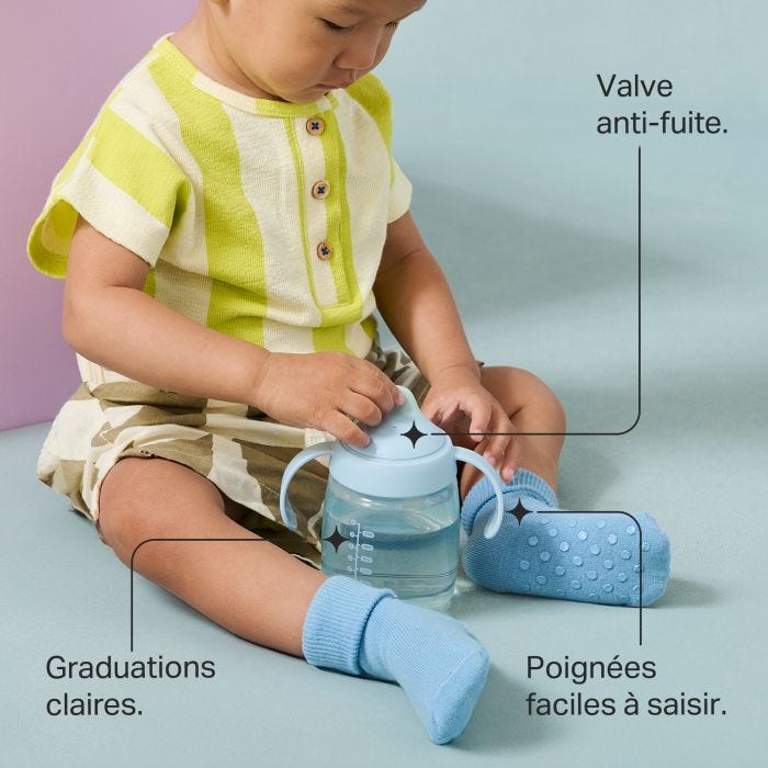 Un enfant en bas âge est assis sur le sol et tient un gobelet à bec d&apos;entraînement avec des indications sur les principales caractéristiques du produit
