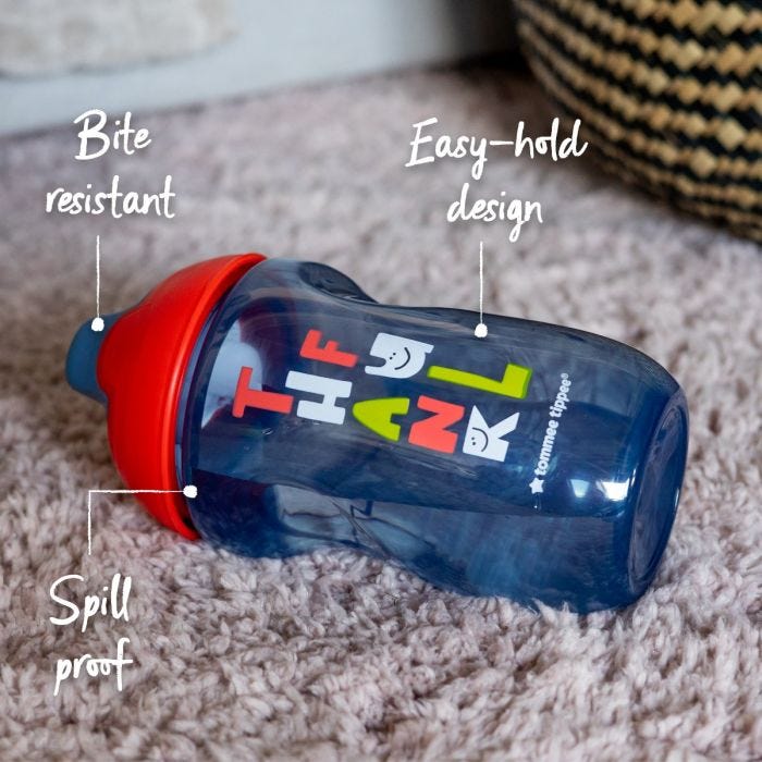 Blue sippee cup lying flat on a carpet with pointers to its bite resistant spout, spill proof lid and easy hold body