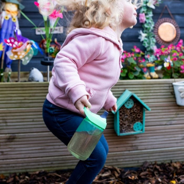 Girl with blonde hair and pink jumper playing in the garden whilst holding the Sportee Water Bottle in Green