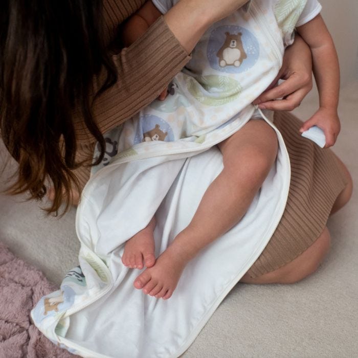 Une femme dans une robe brune assise sur le sol berçant un bébé portant une gigoteuse sleepbag Grofriends dézippée depuis le bas.