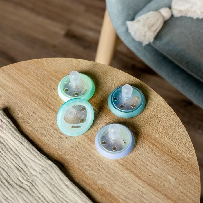 3 Breast-like Night soothers in various colours lying on a brown wooden table.