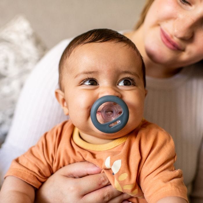 Baby sitting on mum’s knee with blue breast-like pacifier in mouth.