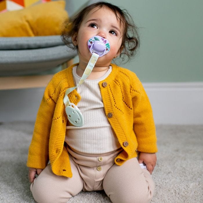 Child sitting on floor with a soother in mouth attached to the soother holder with a green clip.