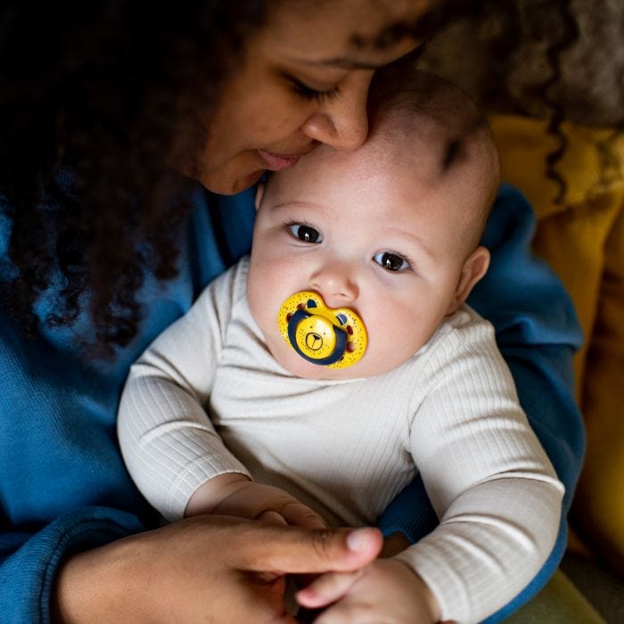 Baby with yellow Fun Style soother in mouth, sitting on mum’s knee and being cuddled by mum.