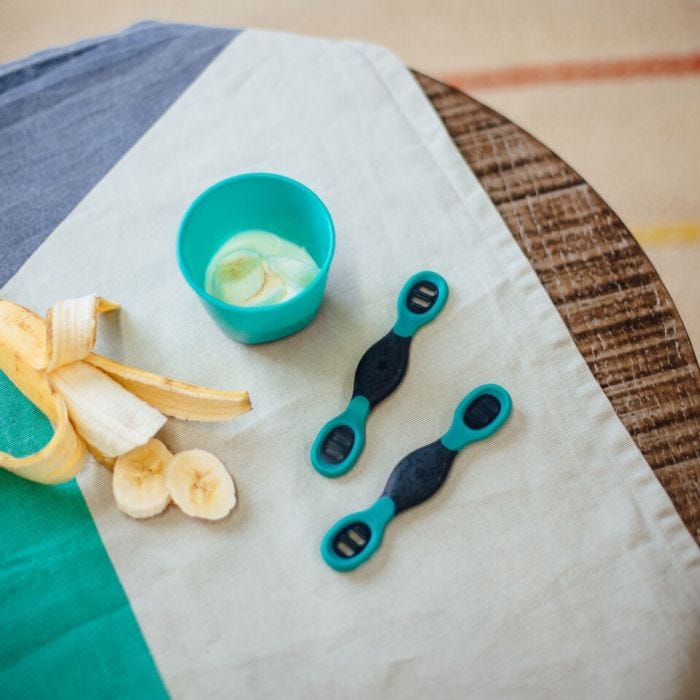 Two Smushee spoons on a kitchen table next to a bananan and bowl of porridge