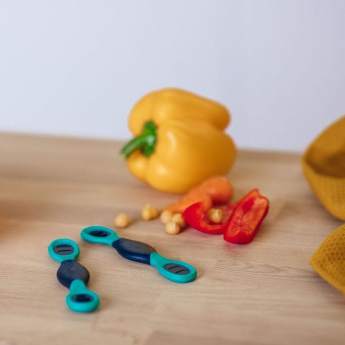 Two Smushee spoons on a kitchen table next to bell peppers