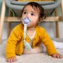 Child sitting on floor with a soother in mouth attached to the soother holder with a purple clip.
