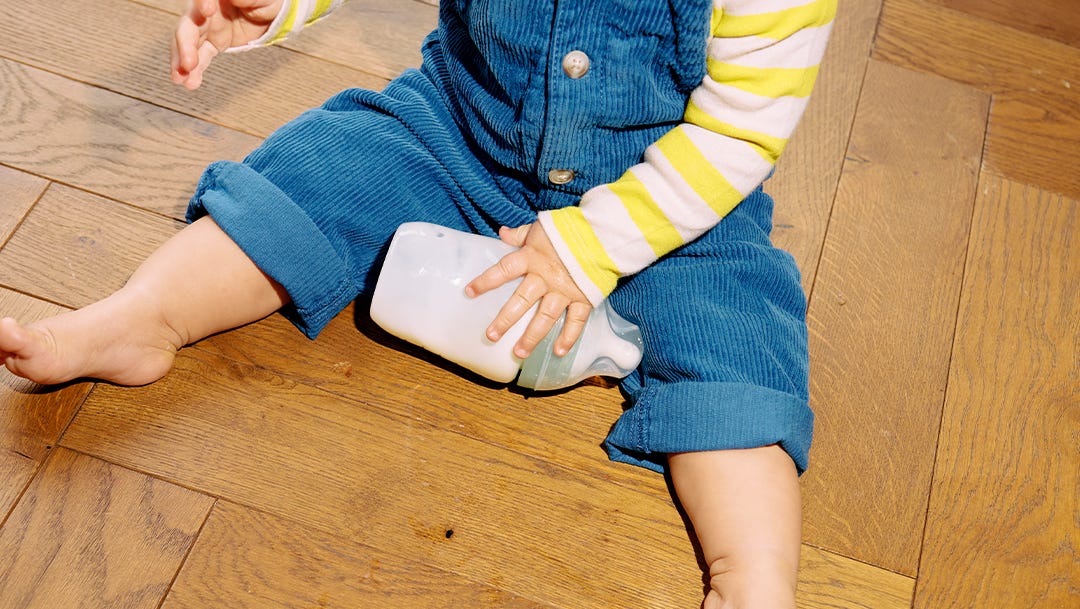 a baby sitting on the floor holding a silicone Tommee Tippee baby bottle 