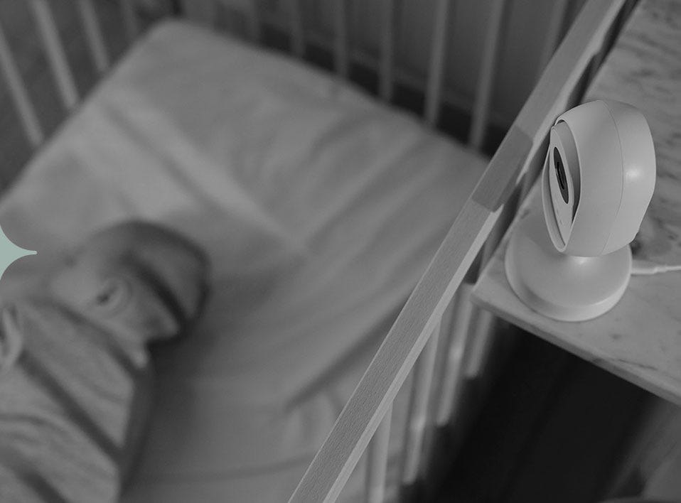 A black and white photo of a baby sleeping in their cot. They're being watched by a sleep monitor.