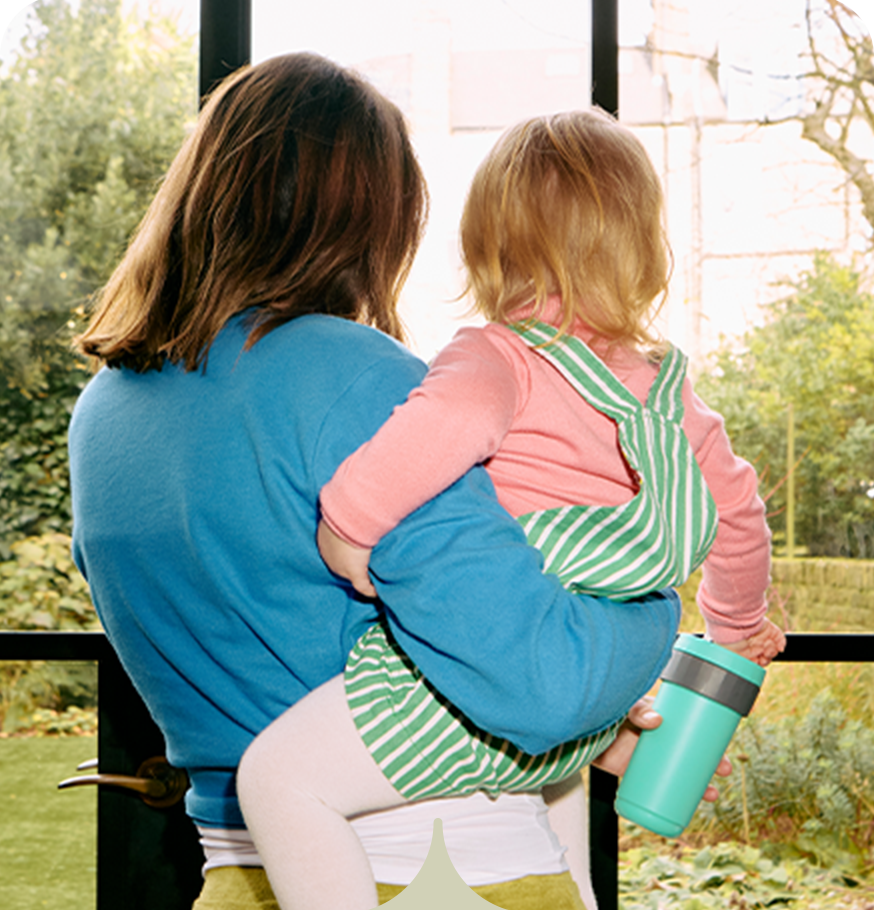 A mum holding her toddler. They are looking out of a window at their garden. 