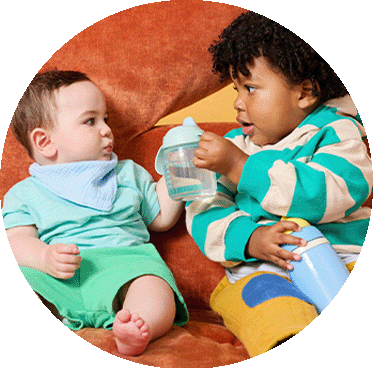 A toddler handing a tommee tippee cup to a younger baby.